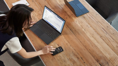 A woman leans over a wooden table, focused on a laptop document, with a smartphone nearby.
