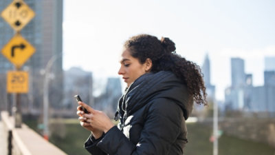 A woman in a black coat looking at her phone near a yellow sign with a black arrow.