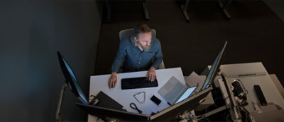 Una persona con la barba è seduta alla scrivania e lavora al computer in una stanza poco illuminata.