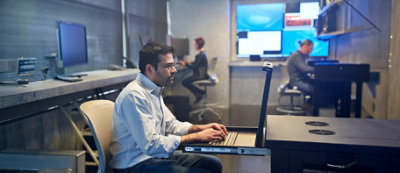 People working at computer stations in an office setting. Man in foreground focused on screen, and woman in background typing on keyboard.