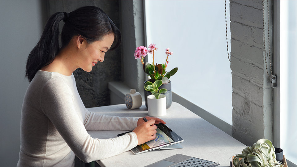 A person at a standing desk smiling while writing with a Surface Slim Pen 2 for Business on a tablet with a Surface Pro Signature keyboard nearby.