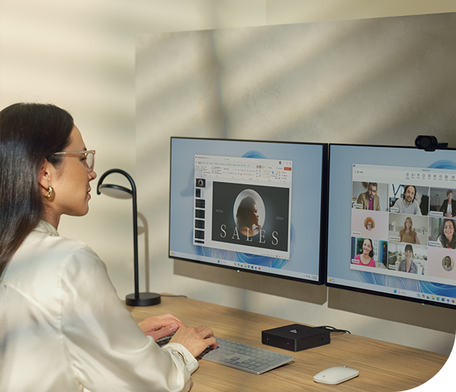 A woman sits at a desk working on two monitors. The left screen displays a sales presentation, while the right screen shows a video conference call with several participants. She is wearing glasses and a white blouse, and the setup includes a keyboard, mouse, and a small desk lamp. The room has soft lighting with shadows from a window.