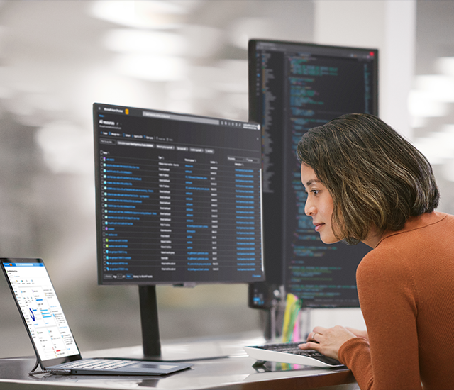 A woman in an orange sweater works at a desk with multiple computer monitors. One monitor shows data tables, another displays code, and a laptop shows graphs. The office setting is modern, with bright, blurred lights in the background, indicating a focus on tasks related to technology or programming. She's focused intently on the screens, suggesting a professional environment.
