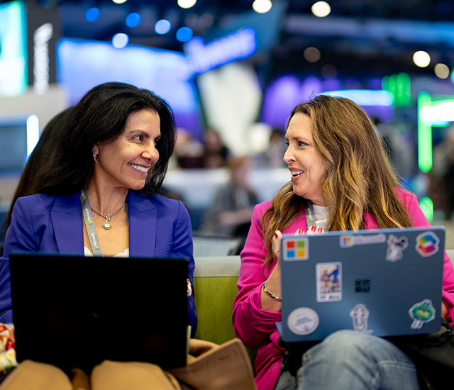 Two women are seated on a green sofa, each with a laptop. The woman on the left wears a blue blazer and looks to the right, smiling. The woman on the right, in a pink jacket, smiles back while holding a laptop with various stickers. They appear to be engaging in a friendly conversation. The background features vibrant, colorful lights and a blurred crowd, suggesting a lively tech event atmosphere.