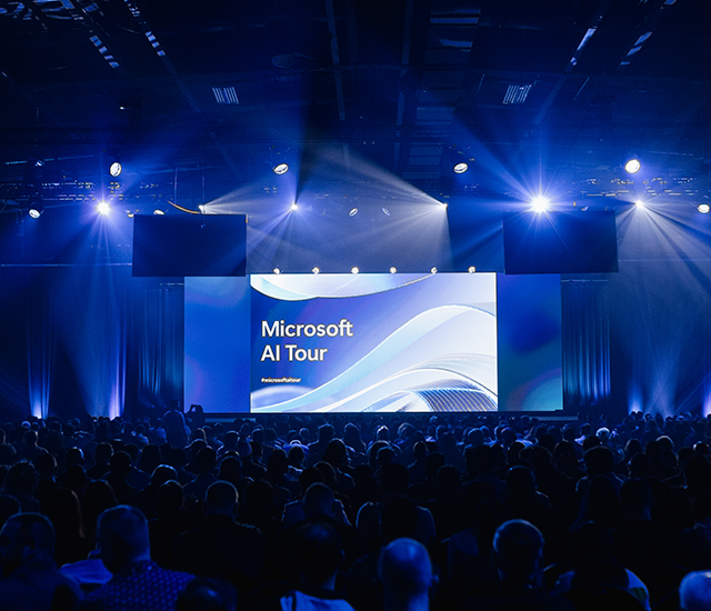 A packed audience at the Microsoft AI Tour event, illuminated by stage lights with a large screen displaying Microsoft AI Tour and a swirling blue and white design. The ambiance is vibrant with attendees facing the illuminated stage, creating a dynamic and engaging atmosphere. Overlayed Text: Microsoft AI Tour microsoftaitour.