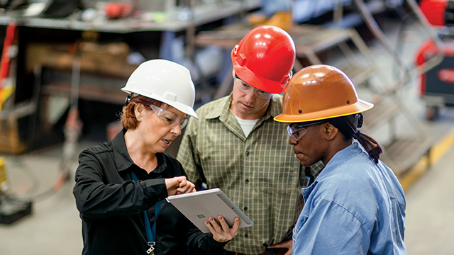 Three workers wearing safety gear and hard hats stand in an industrial environment. A woman with a white hard hat and safety glasses gestures at a tablet, engaging two colleagues: one in a red hard hat and plaid shirt, and another in a brown hard hat and blue shirt. They appear focused, collaborating on tasks amidst blurred background machinery and tools.