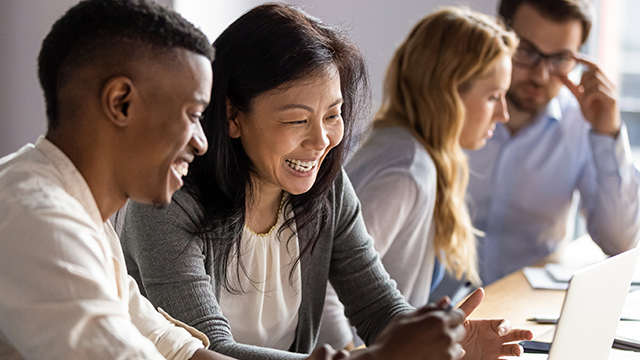 A diverse group of four people sit at a wooden table, engaged in a positive discussion. Two individuals in the foreground are focused on a laptop screen, smiling and gesturing. The background shows two more people, a woman and a man, attentively collaborating. Natural light filters in from the side, creating a bright and welcoming atmosphere conducive to teamwork and collaboration.