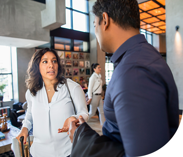 A woman in a white blouse, holding a suitcase, gestures while speaking to a man in a blue shirt in a modern lobby with large windows. A woman walks by in the background. Natural light illuminates the space, showcasing wooden flooring and artwork on the walls.
