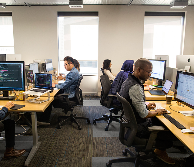 A person in a denim jacket interacts with a tablet in what appears to be a store setting. In the foreground, a soft pink hat and black shoes are on display, suggesting a retail environment. Sunlight streams through a window, creating a casual and professional atmosphere. The scene reflects a blend of technology with traditional shopping experiences.