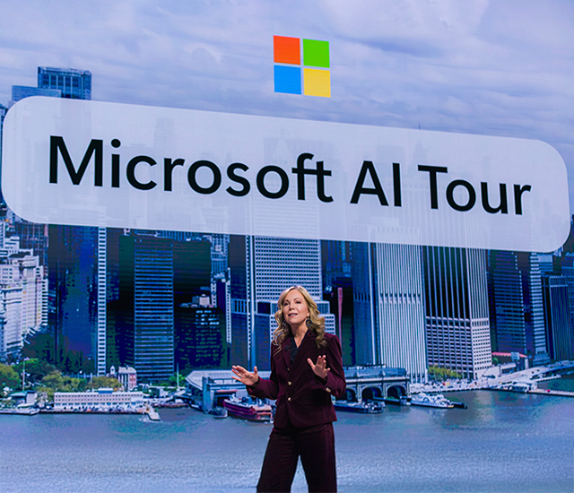 A person with long hair and a beard works at a desk in a dimly lit office. They are using a keyboard and mouse, surrounded by multiple monitors. A laptop, smartphone, glasses, and cables are organized on the desk, creating a tech-focused workspace environment. The setup suggests a professional setting emphasizing technology and multitasking.