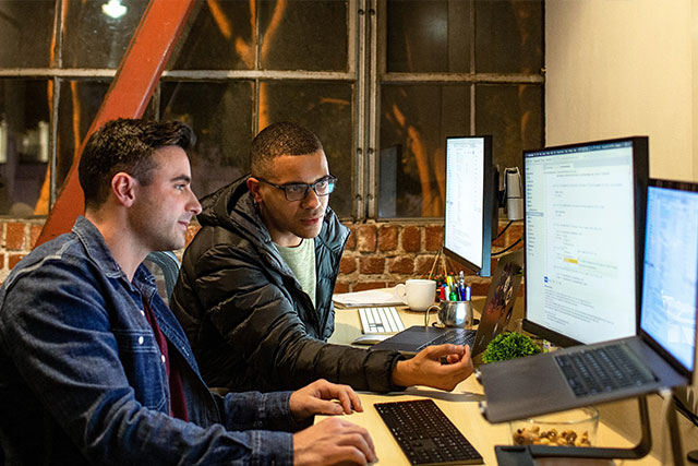 Two people working at desktop computers in an open office space with large windows, viewing software interfaces on dual monitors while sitting at a shared desk.