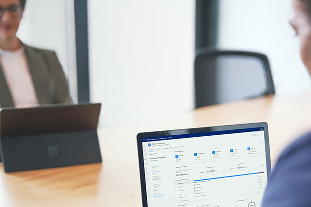 Person using a laptop displaying a financial or data dashboard during a meeting, with another person blurred in the background across a conference table.