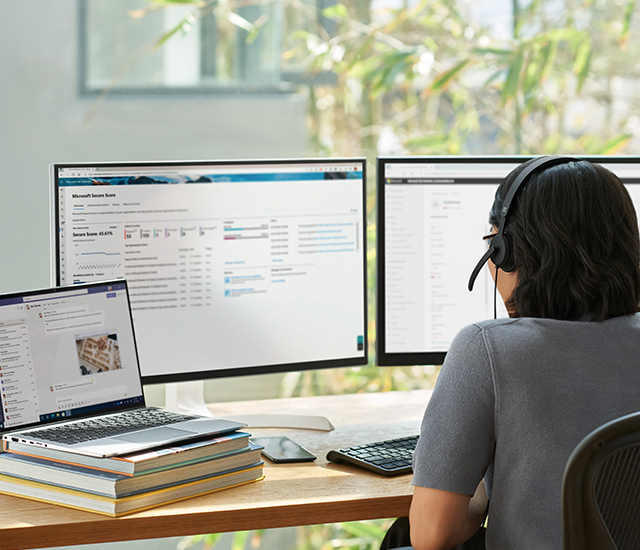 Person wearing a headset working at a desk with two monitors displaying dashboards and a laptop showing a messaging app in a bright home office with plants.