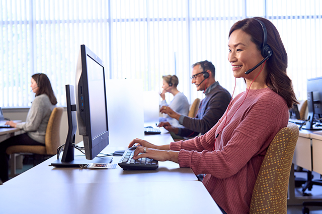 Person wearing a headset sits at a desk typing on a computer in a call center, with several other headset users working at computers in the background.