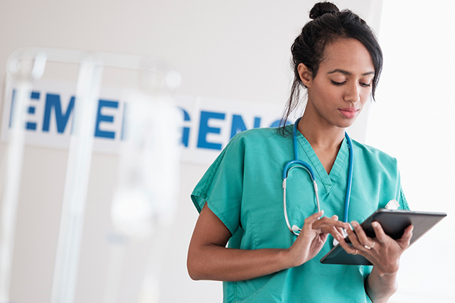 Healthcare professional in scrubs with a stethoscope uses a tablet in a medical setting, with the word “EMERGENCY” blurred in the background.
