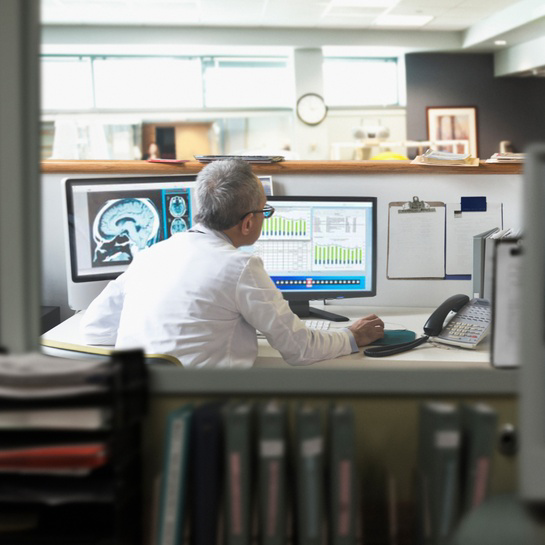 Healthcare professional reviewing medical imaging and data charts on multiple computer monitors at a workstation in a clinical office.