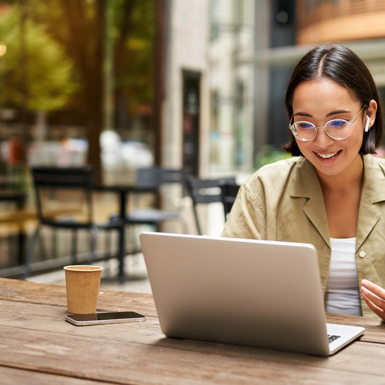 Person using a laptop at an outdoor café table, participating in a virtual meeting with a coffee cup and smartphone nearby.