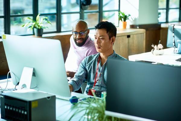 Two people working together at desktop computers in a bright open-plan office with large windows and indoor plants.