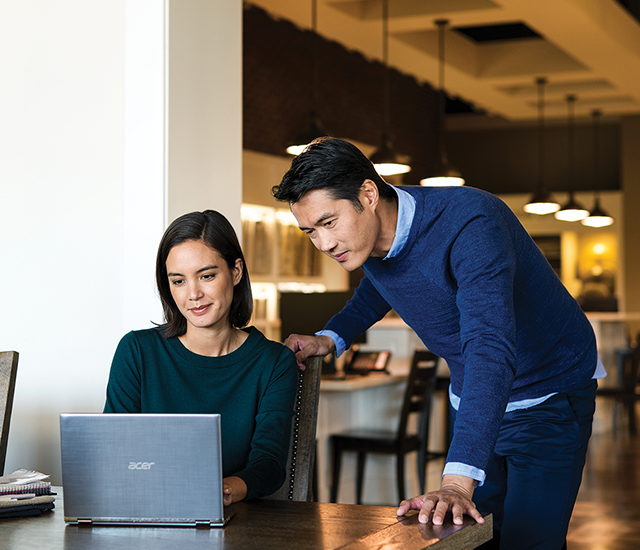 Showroom floor. One person works on a laptop at a desk while another person stands, looking over their shoulder..