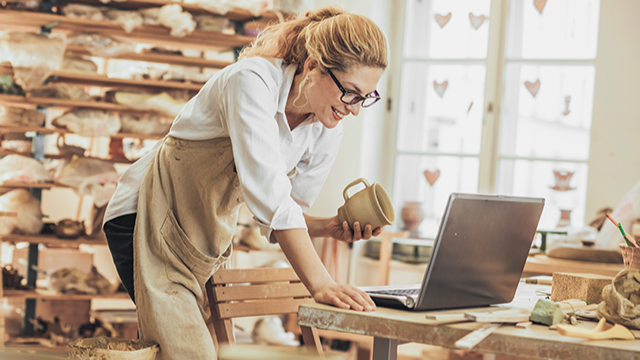 A woman stands in a pottery shop, wearing an apron, while working on a laptop.