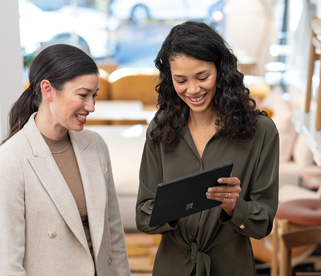 Two women are standing indoors and looking at a tablet. One woman is wearing a beige blazer; the other is in an olive green blouse. They are smiling and appear to be discussing something on the screen..