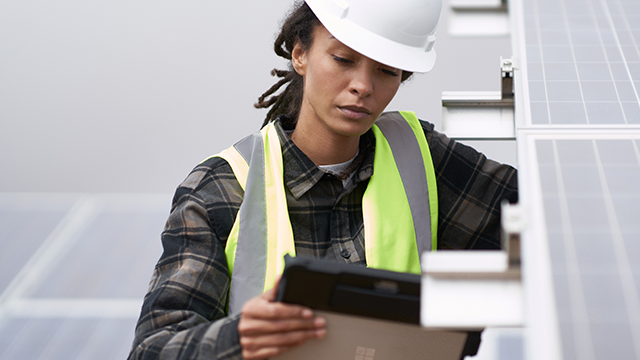 A woman wearing a hard hat and safety vest is holding a tablet, demonstrating her engagement in a construction environment.