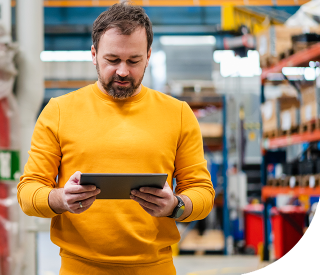 A bearded man in a yellow outfit stands in a warehouse, looking at a tablet. Shelving units and various equipment are visible in the background.