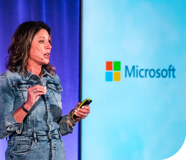A woman wearing a denim jumpsuit speaks on stage while holding a presentation remote. She has a headset microphone and is gesturing with one hand. Behind her is a large screen displaying the Microsoft logo against a light blue background, and the stage is lit with purple lighting.