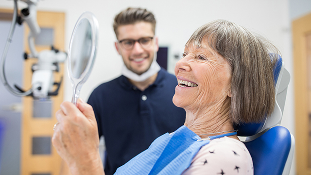 Smiling elderly patient checking her teeth into a hand mirror after her dental treatment. Senior patient checking her teeth after treatment at dental clinic.