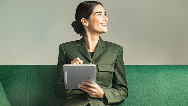 Photograph of a female sitting on a green couch, holding a Surface Pro 12&quot; Platinum device vertically in tablet mode in her left hand, inking with her right hand. She is looking to her left.