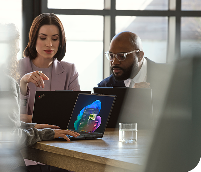 Three professionals engage in a collaborative discussion at a wooden table. Two of them focus intently on a laptop screen showing a vibrant digital interface with a colorful abstract graphic and menu options. One person gestures towards the screen while another watches closely. A glass of water sits nearby, and the room is softly lit through large windows, creating a professional and modern atmosphere.