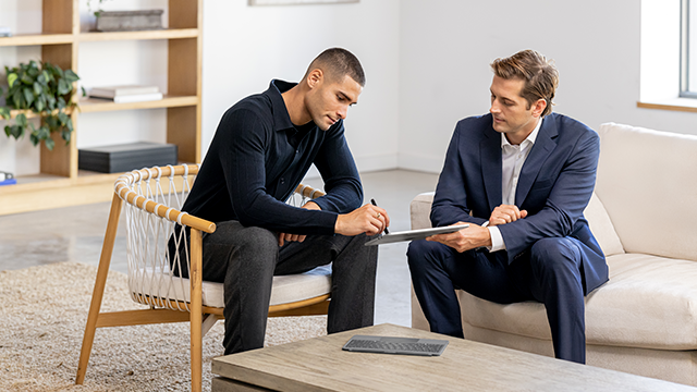 Two men in business attire are seated on a modern beige couch and wooden chair in a contemporary office space. The man on the left, wearing a dark sweater, is taking notes on a tablet, while the man on the right, in a navy suit, gestures toward the tablet. A wooden coffee table is in the foreground, and a bookcase with plants and books is visible in the background, creating a professional atmosphere.