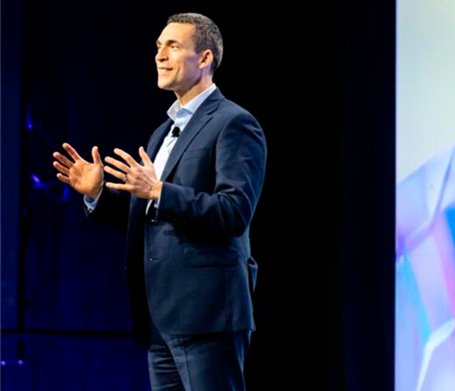 A person in a dark suit and light-colored shirt stands on stage with a microphone clipped to their lapel, engaging in a speech. Their hands are gesturing expressively. The background features a large screen displaying colorful, abstract graphics. The stage is dimly lit, drawing focus to the speaker, who appears confident and poised.