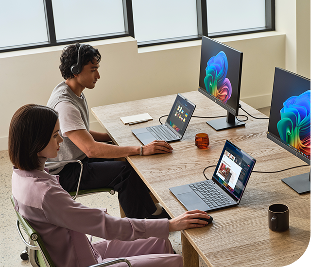 Two individuals work side by side at a wooden desk, each using a laptop connected to desktop monitors showing vibrant, colorful graphics. The person on the left wears a pink outfit and operates a mouse. The person on the right, wearing a t-shirt, listens with headphones. The desk features notebooks, a mug, and a small decorative item, situated in a room with high windows.