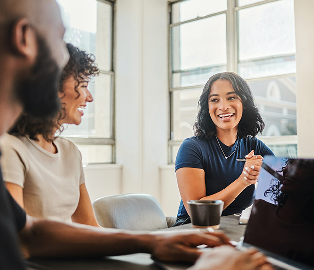 Three people are engaged in a cheerful conversation around a table in a well-lit office with large windows. Two people are visible in profile, while one smiles directly facing them. A laptop and cup are on the table, and natural light fills the room, creating a welcoming and collaborative atmosphere.