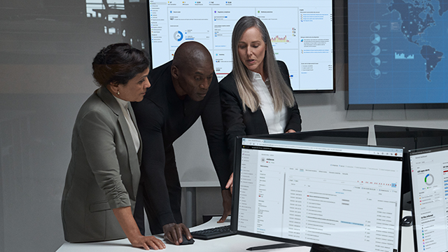 Three professionals stand around a table in a modern office, examining data displayed on computer screens. A woman in a suit points at the screen while another woman and a man attentively follow. A large monitor in the background shows various charts and graphs, indicating a business or analytical context. The scene takes place in a room with large windows, suggesting a professional meeting or collaboration.