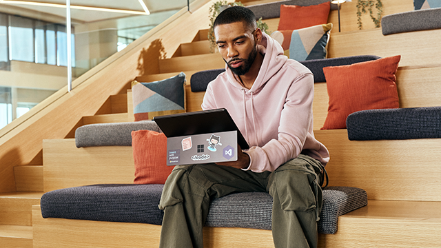 A man in a pink hoodie and green pants sits on a wooden step, using a laptop adorned with various stickers, including and a GitHub Octocat. He is surrounded by colorful cushions in a modern, cozy environment. Steps and natural light add to the relaxed atmosphere, suggesting a casual workspace or study area.