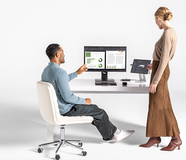 A man seated on a swivel chair points at a computer monitor displaying charts and a document. A woman stands beside him discussing the content. They are at a white desk with a second smaller screen. Both individuals are focused on the information presented, suggesting a collaborative work environment. The minimalistic setting emphasizes the digital content.