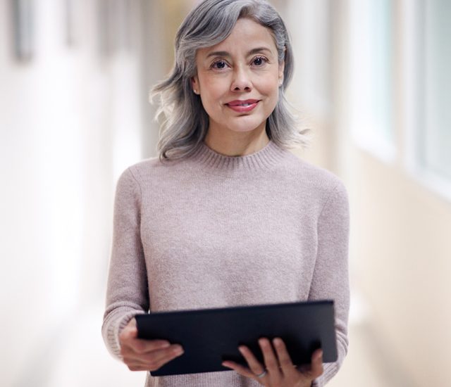 A woman with grey hair, wearing a pink sweater, is standing in a bright, blurred hallway, holding a tablet. She is smiling softly, and her hair falls loosely over her shoulders. The soft lighting creates a warm and inviting atmosphere, highlighting her relaxed and approachable demeanor.