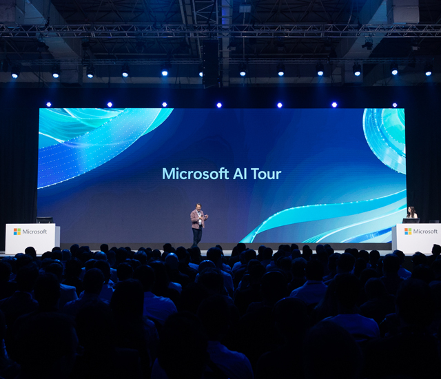 A speaker stands on a stage during the Microsoft AI Tour, addressing a large audience. The backdrop displays vibrant blue digital patterns with Microsoft AI Tour text. Two podiums with the Microsoft logo are on either side of the stage. Overhead lighting creates an engaging atmosphere, emphasizing the event's focus on technology and innovation.