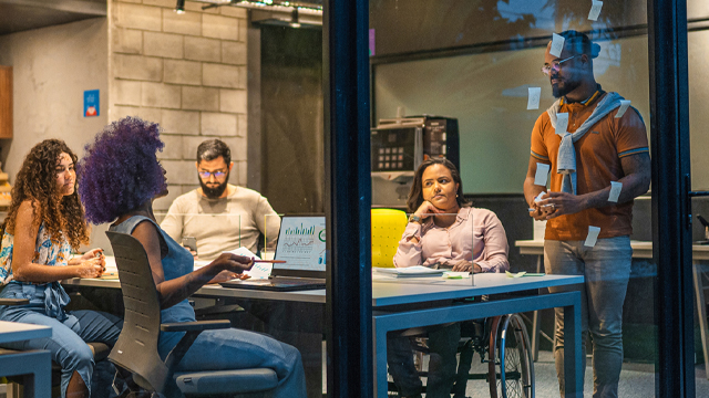 A diverse group of five people in a modern meeting room, engaging in a brainstorming session. One person stands with sticky notes on a glass wall, while others sit attentively, including an individual in a wheelchair. The atmosphere is focused and collaborative, with a laptop and documents on the table. The room has an industrial design with visible concrete walls and ambient lighting.