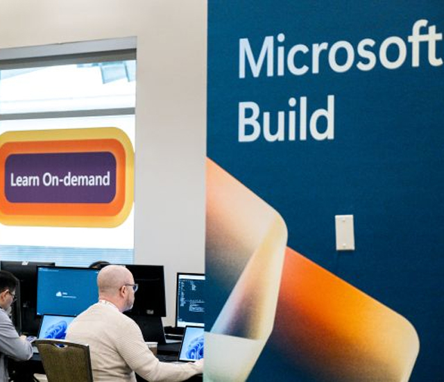A group of people work on laptops and computers in front of a large blue banner that reads Microsoft Build at a tech conference, with a sign in the background that says Learn On-demand.
