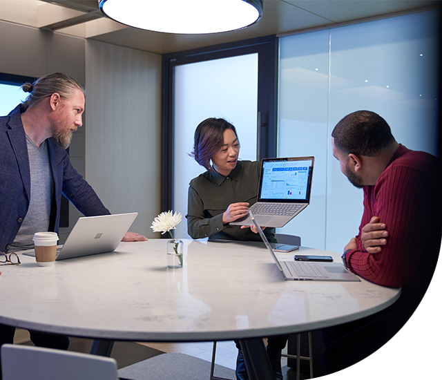 Three people gathered around a round conference table in a meeting room. One person presents analytics and charts displayed on a laptop screen while the others review the information on their own laptops.