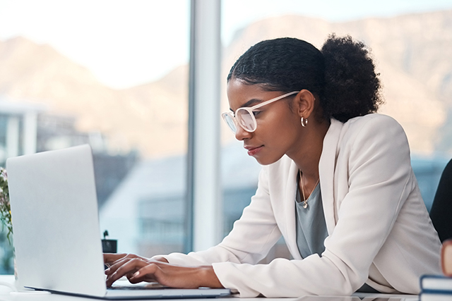 A person wearing glasses and a light-colored blazer working on a laptop at a desk near large windows. A smartphone, books, and a small vase with flowers are visible on the desk, with an urban landscape in the background.