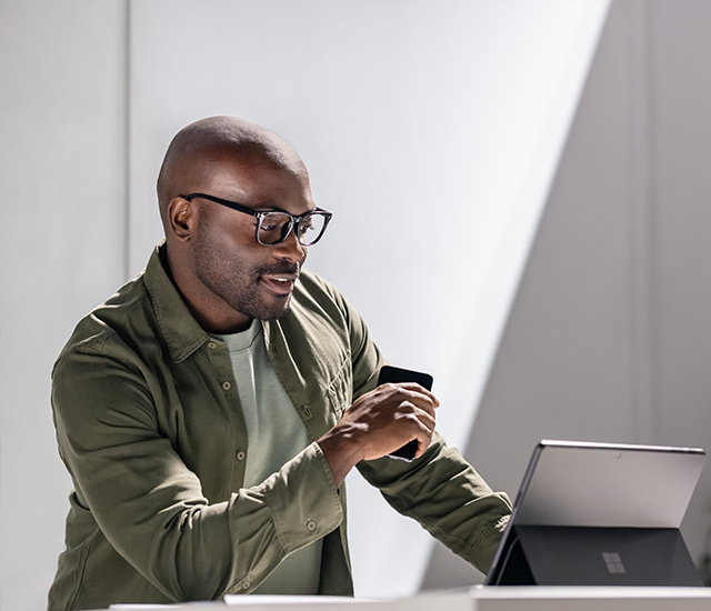 A person wearing glasses and a green button-up shirt stands at a desk using a Microsoft Surface laptop. The person holds a smartphone in one hand while interacting with the laptop. The scene is brightly lit with natural light, featuring a minimalist indoor workspace with a blurred plant in the background. The Microsoft logo is visible on the back of the laptop.
