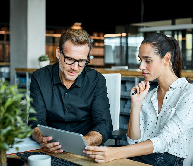 Two colleagues reviewing content on a tablet together at a shared desk in a modern open-plan office.