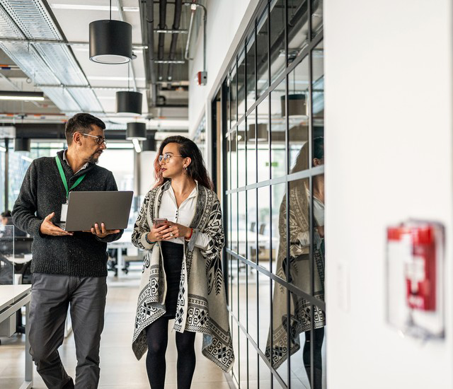 Two coworkers walking and talking in a bright office hallway, one holding an open laptop and the other a coffee cup.