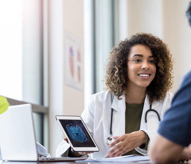 Smiling doctor in a white coat with a stethoscope showing a brain scan on a tablet to a patient during a consultation.