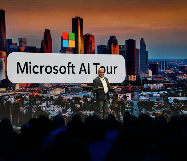 Speaker presenting on stage in front of an audience, with a large screen showing a city skyline and the.