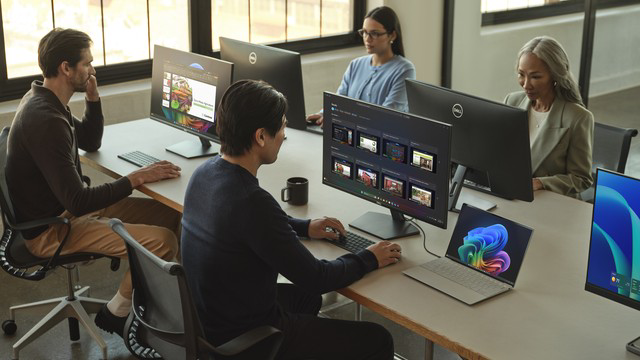 Four colleagues working at desktop monitors and laptops along a shared table in a sunlit office workspace.